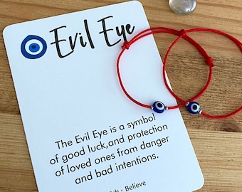 May include: Two red string bracelets with blue evil eye charms. The bracelets are on a white card with the text "Evil Eye" and a description of the meaning of the evil eye symbol.