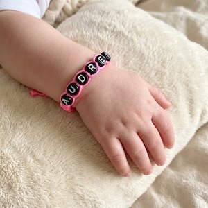 May include: A pink cord bracelet featuring black circular beads with the white letters A, U, D, R, E, Y printed on them. The bracelet is secured with a knot and is worn on a wrist, showcasing the personalised name design in a simple style.