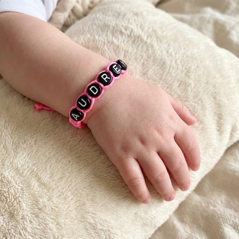 May include: A pink cord bracelet featuring black circular beads with the white letters A, U, D, R, E, Y printed on them. The bracelet is secured with a knot and is worn on a wrist, showcasing the personalised name design in a simple style.