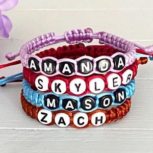 May include: A stack of four adjustable cord bracelets featuring round beads with the names AMANDA, SKYLER, MASON, and ZACH. The bracelets are crafted from lavender, red, light blue, and brown cord, each with white or black letter beads.