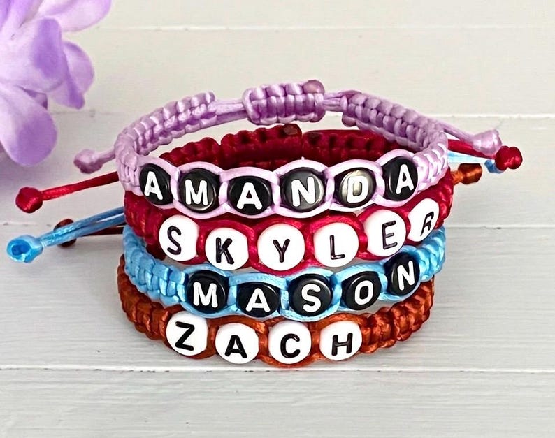 May include: A stack of four adjustable cord bracelets featuring round beads with the names AMANDA, SKYLER, MASON, and ZACH. The bracelets are crafted from lavender, red, light blue, and brown cord, each with white or black letter beads.