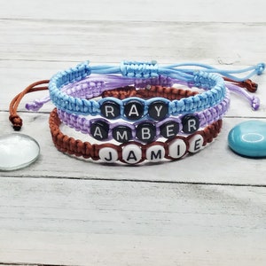 May include: Three adjustable cord bracelets in light blue, lavender, and brown. Each bracelet features round black or white beads with white or black letters spelling out the names RAY, AMBER, and JAMIE, secured with a sliding knot closure.