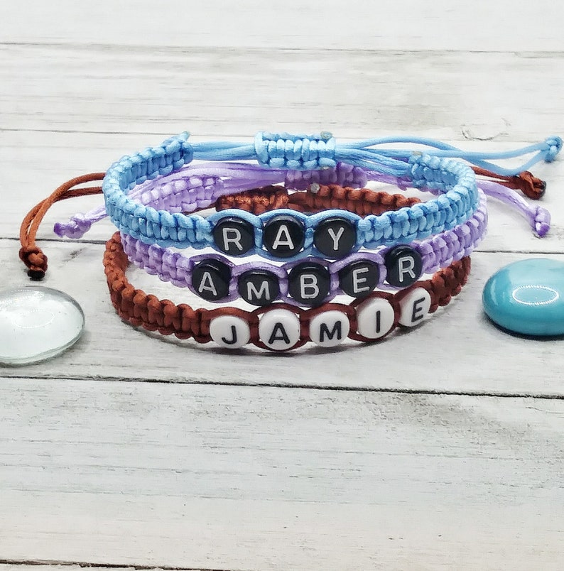 May include: Three adjustable cord bracelets in light blue, lavender, and brown. Each bracelet features round black or white beads with white or black letters spelling out the names RAY, AMBER, and JAMIE, secured with a sliding knot closure.