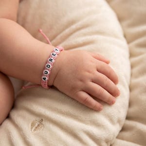 May include: A pink cord bracelet featuring white circular beads with the black printed name AUDREY. The bracelet is constructed with a knotted cord design and includes a thin adjustable string closure, worn around a small wrist.