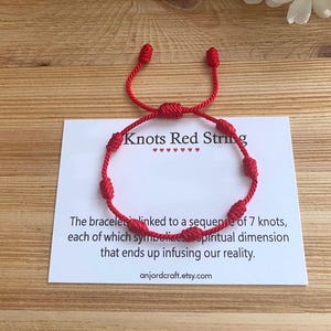 Op de afbeelding: Rood koord armband met zeven knopen. Het armband is gebonden met een slipknoop en heeft een kaart met de tekst "Knots Red String" en "The bracelet is linked to a sequence of 7 knots, each of which symbolizes a spiritual dimension that ends up infusing our reality. anjordcraft.etsy.com".