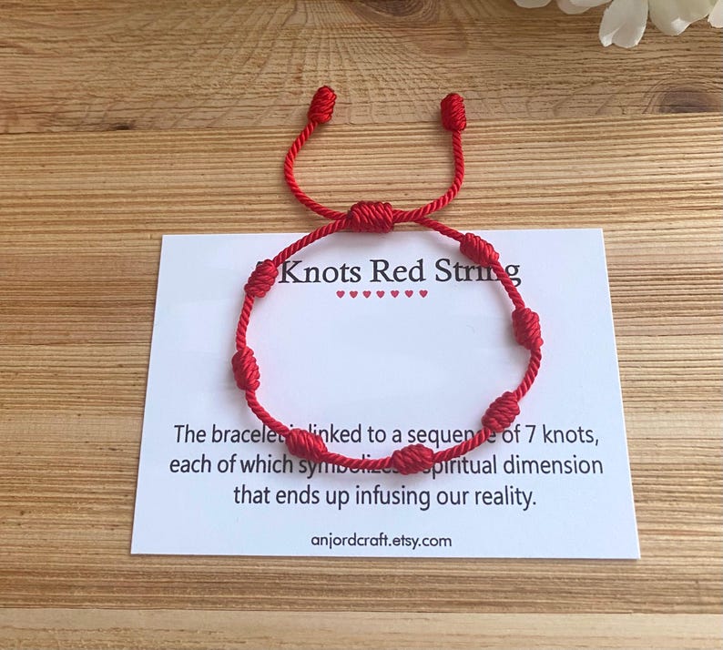 Op de afbeelding: Rood koord armband met zeven knopen. Het armband is gebonden met een slipknoop en heeft een kaart met de tekst "Knots Red String" en "The bracelet is linked to a sequence of 7 knots, each of which symbolizes a spiritual dimension that ends up infusing our reality. anjordcraft.etsy.com".