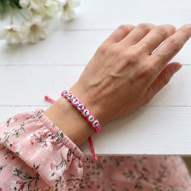 May include: A pink cord bracelet featuring white round beads with black letters spelling out the name NATALIE. The bracelet is adjustable with two thin pink cords extending from the main beaded section, which is worn around the wrist.