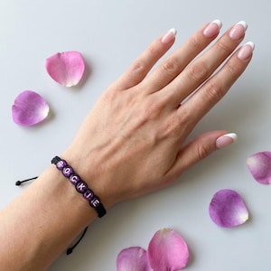 May include: A black cord bracelet featuring purple spherical beads with the white letters R, O, C, K, I, E printed on them. The bracelet has an adjustable sliding knot closure and is displayed on a wrist surrounded by scattered pink rose petals.