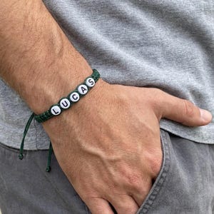 May include: A dark green cord bracelet featuring white round beads with the black printed letters L, U, C, A, and S. The bracelet has adjustable cord ends and is worn around the wrist, showcasing the name Lucas in a simple, clear font style.