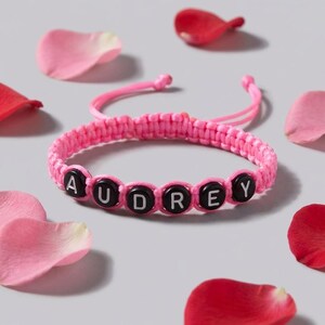 May include: A pink woven cord bracelet featuring six round black beads with the white letters A, U, D, R, E, and Y printed on them. The bracelet has adjustable pink cord ends and is surrounded by scattered pink and red flower petals.