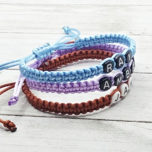May include: Three braided cord bracelets in light blue, lavender, and brown. The light blue bracelet features black beads with the letters R, A, Y. The lavender bracelet has black beads with A, M, B. The brown bracelet has white beads with J, A, M.