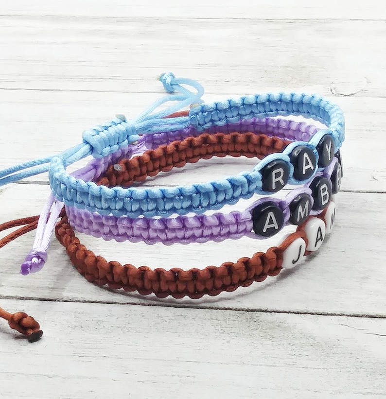 May include: Three braided cord bracelets in light blue, lavender, and brown. The light blue bracelet features black beads with the letters R, A, Y. The lavender bracelet has black beads with A, M, B. The brown bracelet has white beads with J, A, M.
