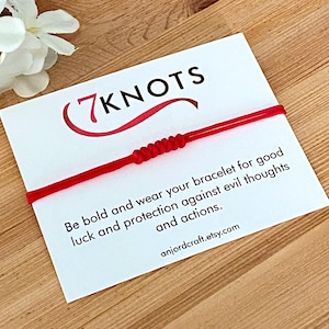 May include: A red string bracelet with a series of knots, displayed on a white card. The card features the text "7 Knots" in black and red, along with the message "Be bold and wear your bracelet for good luck and protection against evil thoughts and actions."