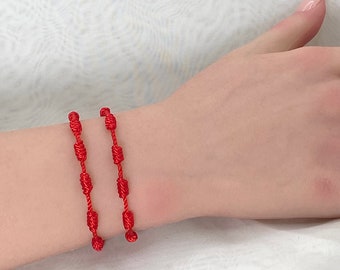 May include: Two red string bracelets with knotted designs. The bracelets are worn on a person's wrist.