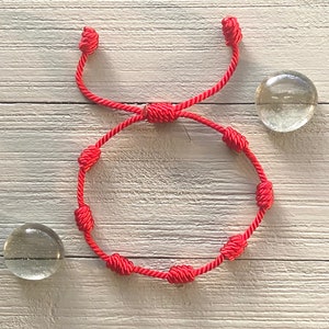 Op de afbeelding: Een rood touwen armband met meerdere knopen. De armband ligt op een wit houten oppervlak.