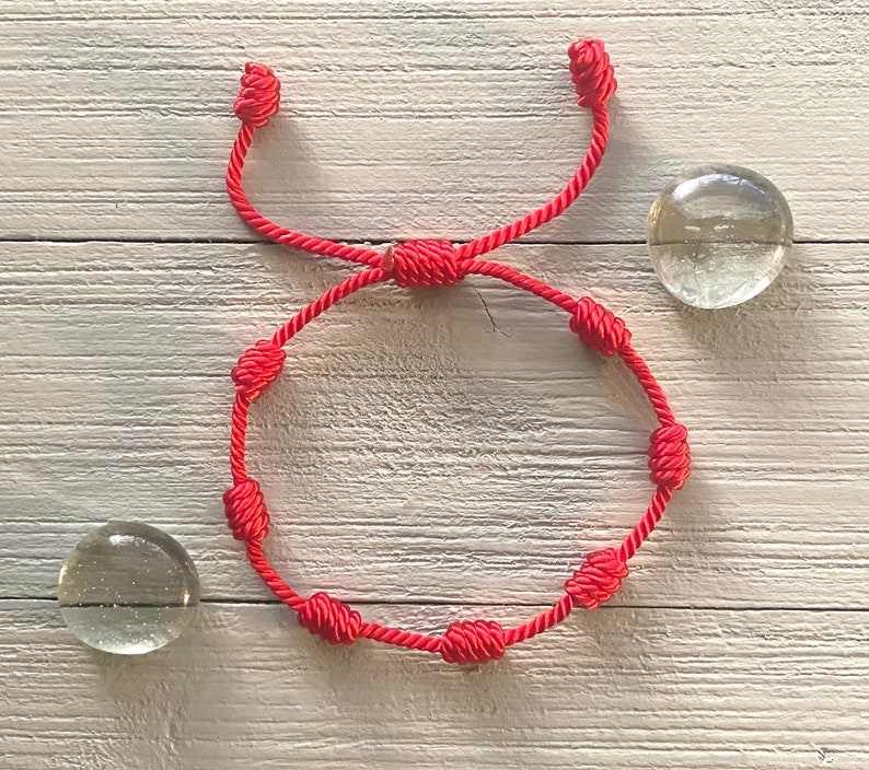 Op de afbeelding: Een rood touwen armband met meerdere knopen. De armband ligt op een wit houten oppervlak.
