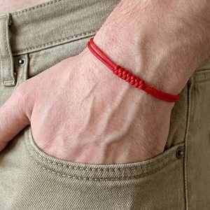 May include: A red woven bracelet is worn on a wrist, with a knot in the center. The bracelet is made of two strands of red cord. The person is wearing khaki-colored pants.