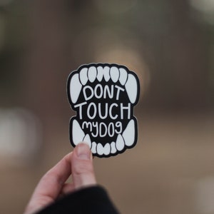 May include: A black and white sticker with the text "DON'T TOUCH MY DOG" in a stylized font. The sticker is shaped like a set of teeth and is held up by a hand.