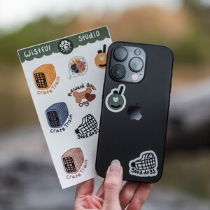 May include: A black smartphone with multiple stickers, held next to a sheet of stickers. The stickers feature illustrations of dog crates, a dog bone, and the words "Crate Train" and "Good Dogs." The sheet is labeled "Wistful Studio."