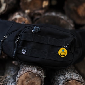 Könnte beinhalten: Schwarze Bauchtasche mit Katzenlogo und gelber Anstecknadel mit der Aufschrift "Ausgeglichener Hundehalter" auf einem Stapel Holzscheite.