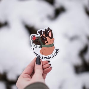 May include: A white sticker with a cartoon dog wearing a green collar and antlers. The dog is holding a shopping basket with a red apple inside. The text "muzzled misfit club" is written in black below the dog.