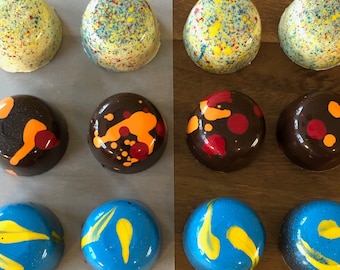 May include: An assortment of twelve artisan chocolates. The chocolates are arranged in three rows of four. The top row has yellow and blue speckled chocolates. The middle row has dark chocolate with orange and red designs. The bottom row has blue and yellow swirl designs.