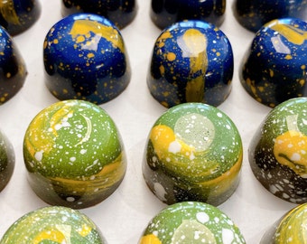 Puede incluir: Una vista cenital de una colección de bombones artesanales. Los bombones tienen forma de cúpula y un acabado brillante. Algunos son de color azul oscuro con salpicaduras doradas, mientras que otros son verdes con detalles amarillos y blancos.