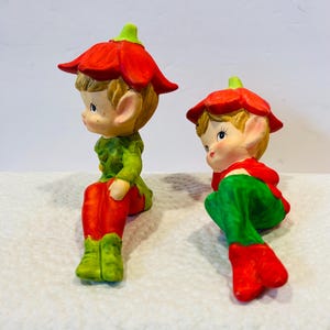 May include: Two ceramic elf figurines with red and green clothing. The elves are wearing red flower hats and have painted facial features.