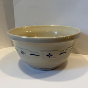 Puede incluir: Un cuenco de cerámica color crema con un borde ancho y una base redondeada. El exterior presenta un patrón de banda en relieve repetitivo decorado con pequeños motivos florales azul oscuro y líneas onduladas horizontales.