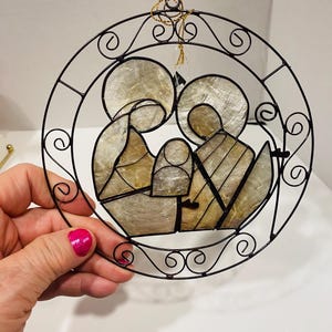 Può includere: Un ornamento natalizio rotondo in metallo nero raffigurante la Sacra Famiglia. Le figure sono riempite con un materiale perlescente e l'ornamento ha un cordino dorato per appenderlo. Il design presenta una cornice circolare con volute decorative.