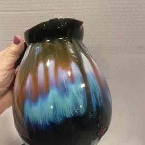 May include: A handcrafted ceramic vase with a unique, flowing glaze. The vase features a dark, glossy rim that transitions into a blend of brown, white, blue, and teal hues, creating a striking visual effect. The colors cascade down the vase, adding depth and artistic flair.