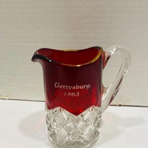 May include: A small, ruby-red glass pitcher with a clear handle and base. The rim is trimmed with gold. The words "Gettysburg 1863" are etched in white on the side. The base has a diamond-cut pattern.