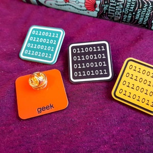 May include: Four enamel pins with a binary code design. The pins are square shaped and have a black, yellow, green, and orange background. The text on the pins reads "01100111 01100101 01100101 01101011". One of the pins has the word "geek" written on it.