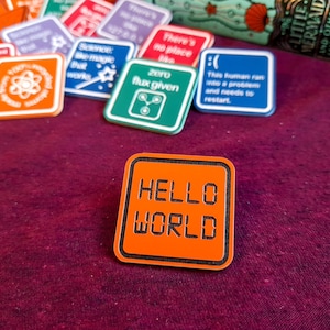 May include: An orange square pin with a black border and the text "HELLO WORLD" in black.