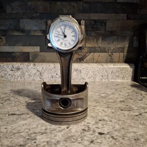 Peut inclure: Une horloge fabriquée à partir d'un piston de moteur de voiture. Le cadran de l'horloge est blanc avec des chiffres romains noirs et des aiguilles noires. Le piston est argenté et a un anneau noir autour du haut.