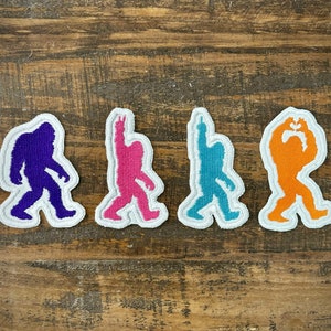 May include: Four embroidered iron-on patches featuring different colored silhouettes of Bigfoot. The patches are white with a black outline. The Bigfoot are purple, pink, blue, and orange.