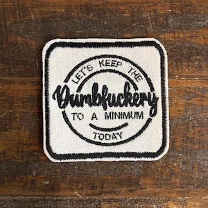 May include: Square patch with a white background and black border. The patch features the text "LET'S KEEP THE Dumbfuckery TO A MINIMUM TODAY" in a circular design. The patch is likely an iron-on or sew-on.