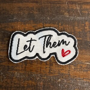 May include: Embroidered patch with the words "Let Them" in black script, a small red heart, and a black border. The patch is white and has a cloud-like shape. The patch is on a wooden surface.