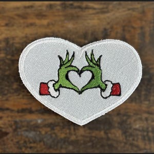 May include: White heart-shaped patch with green hands making a heart shape. The hands are wearing red and white cuffs.
