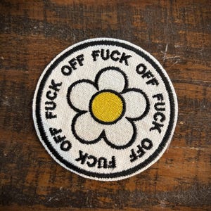 May include: Embroidered patch with a white daisy design and a yellow center. The patch is circular with the words "FUCK OFF" in black, repeated around the edge.