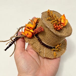 May include: A small top hat crafted from tan burlap fabric, featuring a dark brown trim around the base. The hat is decorated with orange fabric flowers, small clusters of orange berries, and thin, dangling brown twine strings.