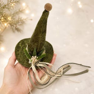 May include: A pointed olive green velvet hat featuring a small brown pom-pom at the tip. The base is decorated with a cluster of white berries, green pine needles, and several long, trailing ribbons in shades of cream, olive, and dark green.