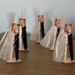 May include: Set of five wooden clothespin wedding favors. Each clothespin is painted to look like a bride and groom kissing. The clothespins are decorated with white tulle and a silver pin.