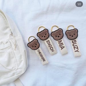 May include: Four keychains featuring a brown, fuzzy, embroidered bear head attached to a cream-colored fabric strap. Each strap is embroidered with a name in brown thread: THEODORE, ENOCH, MIA, and SUZY. Each bear head has a gold-toned metal ring.