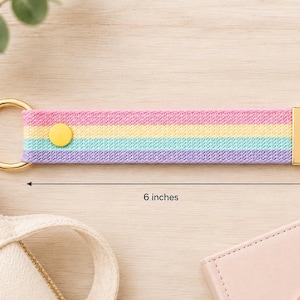 May include: A fabric keychain featuring horizontal stripes in pink, yellow, light blue, and lavender. It includes a gold-tone metal ring, a yellow snap button, and a gold-tone end cap. The keychain measures 6 inches in length and 1 inch in width.
