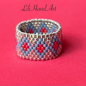 Op de afbeelding: Een kralenring met een zilveren band en een patroon van blauwe en rode kralen. De ring staat op een roze achtergrond. De tekst "Lili Hand Art" staat in het midden van de afbeelding.