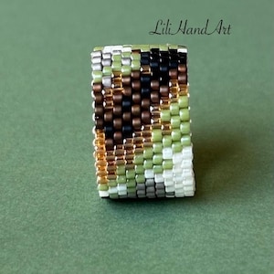 May include: A beaded ring with a rectangular shape. The ring features a pattern of small, colorful beads in shades of green, brown, black, gold, white, and gray. The text "Lili Hand Art" is visible in the upper right corner. The ring is set against a green background.