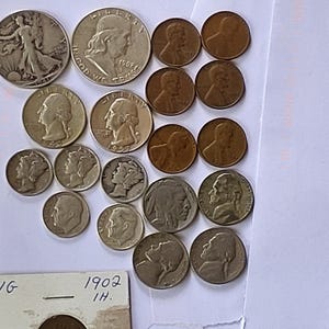 May include: A collection of vintage coins, including silver half dollars, quarters, dimes, nickels, and pennies. The coins are arranged on a white surface, showcasing various designs and dates. A coin holder with the text "1902" is also visible.