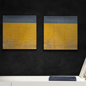 May include: Two abstract paintings with a yellow and gray color scheme hang on a black wall. A black lamp, books, a white cat figurine, and a clear glass candle holder are on a white shelf. The paintings have a textured appearance.