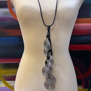 May include: A long black leather necklace with a silver pendant featuring multiple concentric circles. The pendant hangs from a tassel of black leather cords.